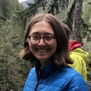 Smiling headshot of a person with light skin, chin-length brown hair and glasses, wearing a blue jacket and standing in a conifer forest